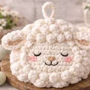 Puede incluir: Un salvamanteles con forma de cordero tejido a ganchillo, de color crema, con un lazo para colgar. El cordero tiene los ojos cerrados, mejillas rosas y orejas internas de color canela. Se encuentra sobre una tabla de cortar de madera, con huevos de Pascua en el fondo.