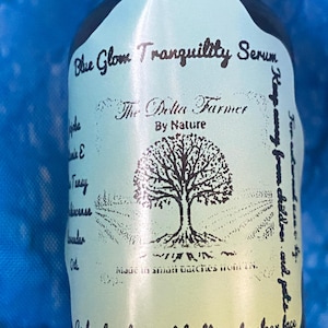 May include: A brown glass bottle with a light blue label featuring the text "Blue Glow Tranquility Serum" and "The Delta Farmer By Nature." The label includes a tree graphic and text about ingredients and usage. The bottle is against a blue background.