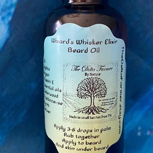 May include: A dark glass bottle of Wizard's Whisker Elixir Beard Oil. The label is light green with black text and a tree illustration. The text includes instructions for use and the name of the maker, The Delta Farmer.