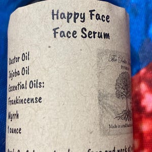 May include: A brown glass bottle with a beige textured label featuring the text Happy Face Face Serum. The label lists ingredients including Castor Oil, Jojoba Oil, and essential oils like Frankincense and Myrrh, with a volume of 1 ounce.