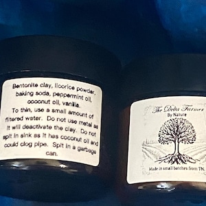 May include: Two small, dark jars with white labels. One label lists ingredients: bentonite clay, licorice powder, baking soda, peppermint oil, coconut oil, and vanilla. The other label reads "The Delta Farmer By Nature" with a tree graphic.
