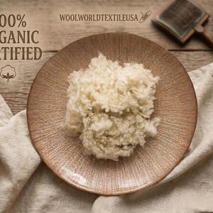 May include: A pile of raw, off-white organic wool fibers centered on a textured, circular bronze-colored plate. The text 100% ORGANIC CERTIFIED and WOOLWORLDTEXTILEUSA are visible, along with a small cotton icon, a hand carder, and a drop spindle.