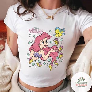 Może przedstawiać: Biały t-shirt z kr&oacute;tkim rękawem z nadrukiem graficznym przedstawiającym Arielkę z Małej Syrenki z długimi r&oacute;żowymi włosami, fioletowym topem z muszelek i ż&oacute;łtą rybką Florkiem, w otoczeniu małych konik&oacute;w morskich i bąbelk&oacute;w z tytułem filmu.