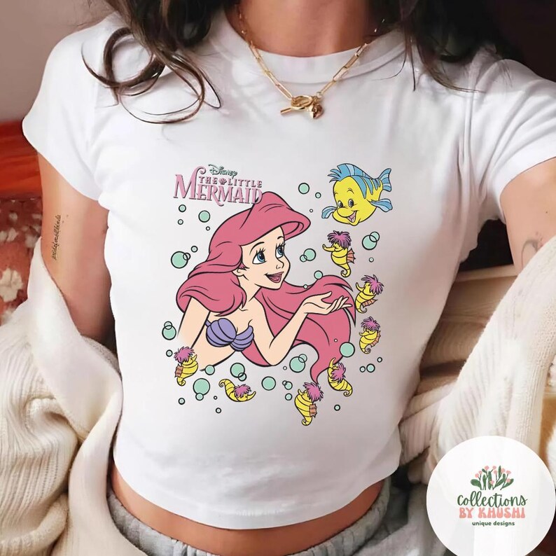 Może przedstawiać: Biały t-shirt z kr&oacute;tkim rękawem z nadrukiem graficznym przedstawiającym Arielkę z Małej Syrenki z długimi r&oacute;żowymi włosami, fioletowym topem z muszelek i ż&oacute;łtą rybką Florkiem, w otoczeniu małych konik&oacute;w morskich i bąbelk&oacute;w z tytułem filmu.