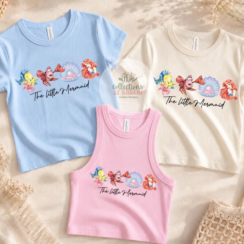 May include: A light blue t-shirt, a cream t-shirt, and a pink tank top, each featuring a graphic of Flounder, Sebastian, a pearl in a shell, and Ariel, with the text The Little Mermaid printed in black script below the characters.