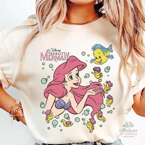 Może przedstawiać: Kremowy t-shirt z nadrukiem graficznym przedstawiającym Arielkę z Małej Syrenki z długimi r&oacute;żowymi włosami, fioletowym topem z muszelek oraz niebiesko-ż&oacute;łtą rybką Florkiem. Wz&oacute;r zawiera kilka małych ż&oacute;łtych konik&oacute;w morskich i bąbelki.