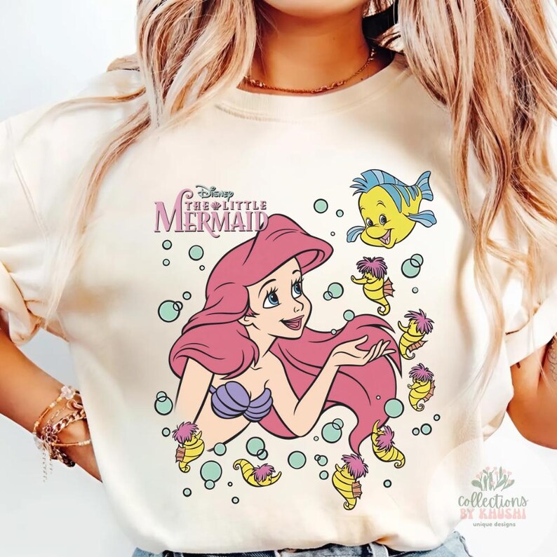 Może przedstawiać: Kremowy t-shirt z nadrukiem graficznym przedstawiającym Arielkę z Małej Syrenki z długimi r&oacute;żowymi włosami, fioletowym topem z muszelek oraz niebiesko-ż&oacute;łtą rybką Florkiem. Wz&oacute;r zawiera kilka małych ż&oacute;łtych konik&oacute;w morskich i bąbelki.