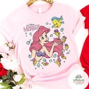 Może przedstawiać: Jasnor&oacute;żowy t-shirt z grafiką przedstawiającą Arielkę z Małej Syrenki Disneya, otoczoną przez Florka, kilka konik&oacute;w morskich i bąbelki. Napis "Disney THE LITTLE MERMAID" jest nadrukowany na fioletowo nad ilustracją postaci.