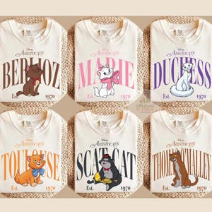 May include: Six off-white t-shirts featuring Disney The Aristocats characters: Berlioz, Marie, Duchess, Toulouse, Scat Cat, and Thomas O'Malley. Each shirt displays the character's name in large letters, the text Est. 1970, and the Disney logo.