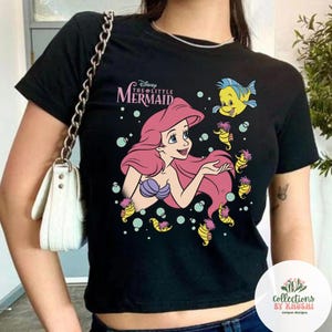 Może przedstawiać: Czarny t-shirt z kr&oacute;tkim rękawem z nadrukiem graficznym przedstawiającym Arielkę z Małej Syrenki z długimi r&oacute;żowymi włosami, fioletowym topem z muszelek, ż&oacute;łtymi konikami morskimi i bąbelkami. Powyżej nadrukowano tekst Disney The Little Mermaid.