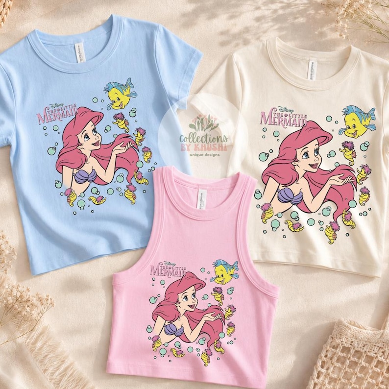 Może przedstawiać: Trzy koszulki z grafiką z Małej Syrenki Disneya. Jasnoniebieski t-shirt z kr&oacute;tkim rękawem, kremowy t-shirt z kr&oacute;tkim rękawem oraz r&oacute;żowy bezrękawnik. Każda koszulka przedstawia Arielkę, Florka i koniki morskie w otoczeniu baniek.