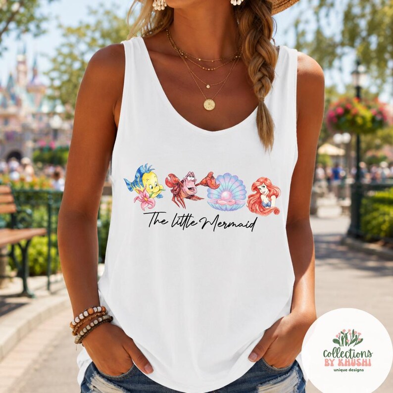 May include: A white sleeveless tank top featuring a graphic print of Flounder, Sebastian, a pearl in a shell, and Ariel, with the text The Little Mermaid written in black script below the characters. The top has a scoop neckline and wide straps.