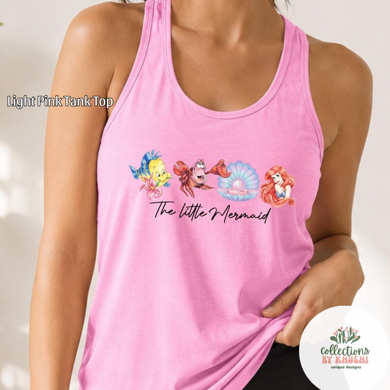 May include: A light pink racerback tank top featuring a graphic print of Flounder, Sebastian, a pearl in a shell, and Ariel. The text "The Little Mermaid" is printed in black script below the characters. The words "Light Pink Tank Top" appear.