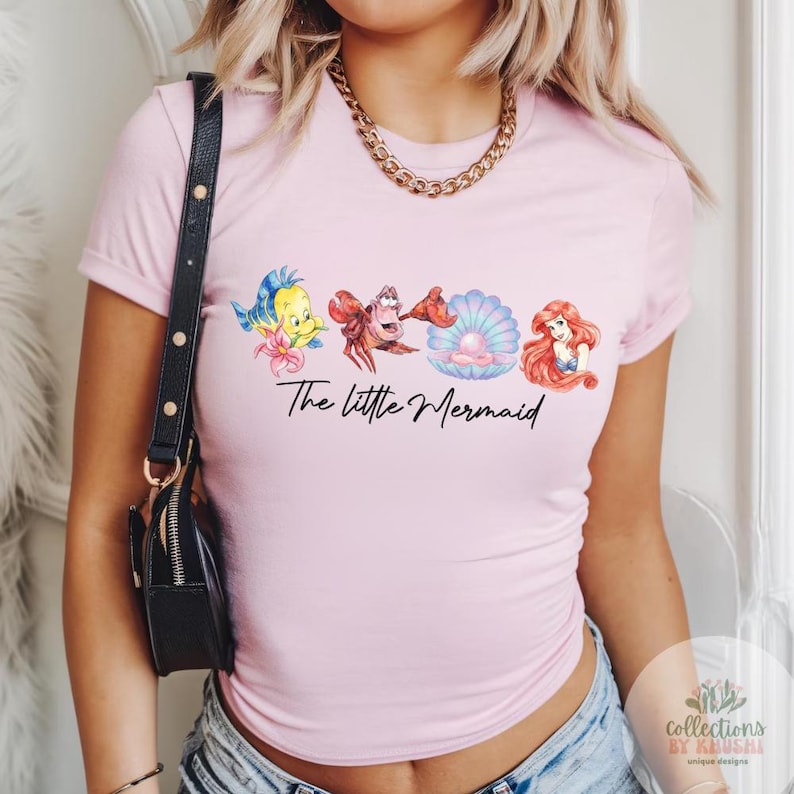 May include: A light pink short-sleeve t-shirt featuring a graphic print of Flounder, Sebastian, a pearl in a shell, and Ariel. The text The Little Mermaid is printed in black script below the characters. A gold chain necklace is worn around the neck.