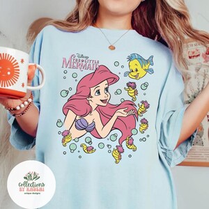 Może przedstawiać: Jasnoniebieski t-shirt z nadrukiem graficznym przedstawiającym Arielkę z Małej Syrenki z długimi r&oacute;żowymi włosami, fioletowym topem z muszli oraz Florka. Wz&oacute;r zawiera małe koniki morskie i bąbelki oraz nadrukowany powyżej napis Disney The Little Mermaid.