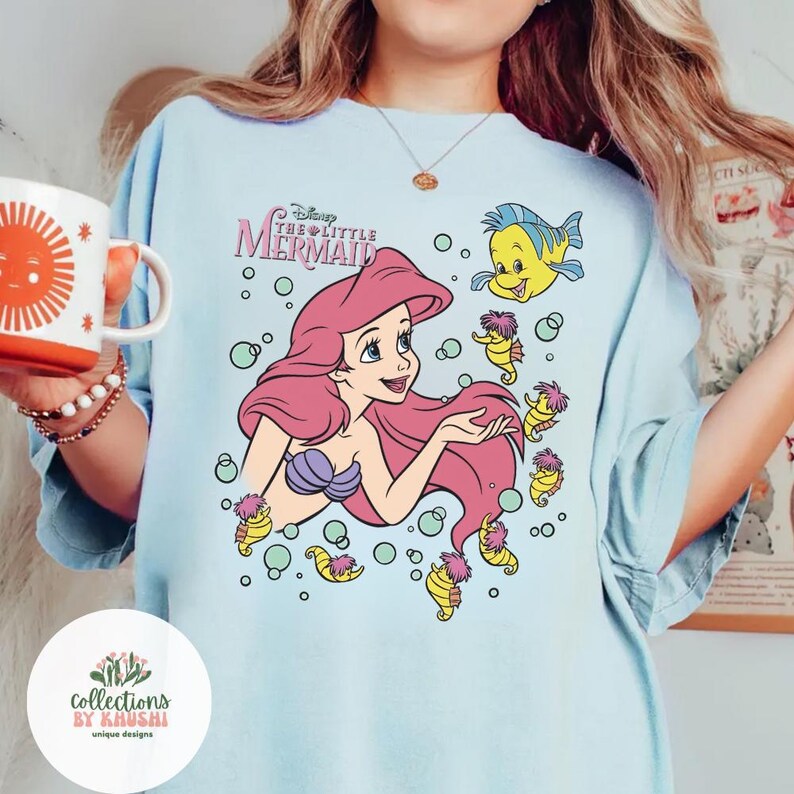 Może przedstawiać: Jasnoniebieski t-shirt z nadrukiem graficznym przedstawiającym Arielkę z Małej Syrenki z długimi r&oacute;żowymi włosami, fioletowym topem z muszli oraz Florka. Wz&oacute;r zawiera małe koniki morskie i bąbelki oraz nadrukowany powyżej napis Disney The Little Mermaid.