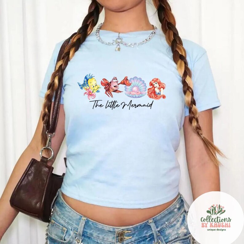May include: A light blue short-sleeve t-shirt featuring a graphic print of Flounder, Sebastian, a pearl in a shell, and Ariel. The text "The little Mermaid" is printed in black script below the characters. The shirt has a crew neckline.