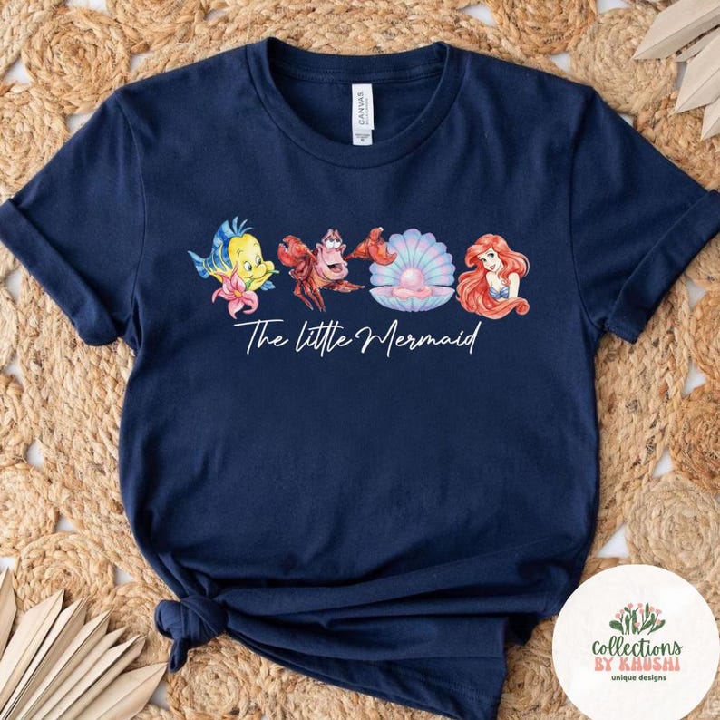 May include: A navy blue short-sleeve t-shirt featuring a graphic print of Flounder, Sebastian, a pearl in a shell, and Ariel. Below the characters, the text reads "The Little Mermaid" in a white script font. The shirt has a crew neckline.