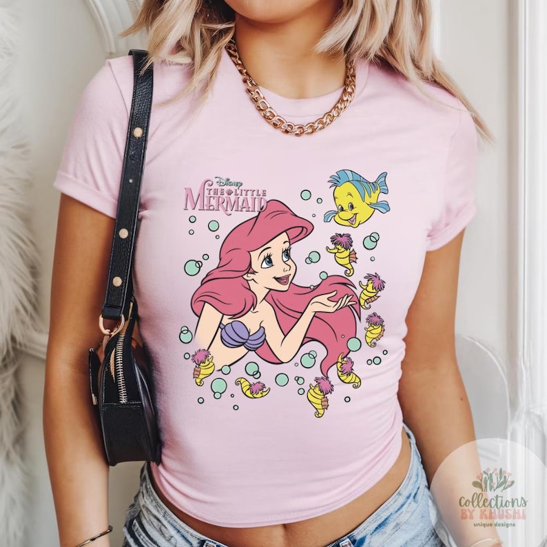 Może przedstawiać: Jasnor&oacute;żowy t-shirt z kr&oacute;tkim rękawem z nadrukiem graficznym przedstawiającym Arielkę z Małej Syrenki, Florka oraz kilka konik&oacute;w morskich otoczonych bąbelkami. Nad ilustracją postaci znajduje się napis Disney The Little Mermaid.