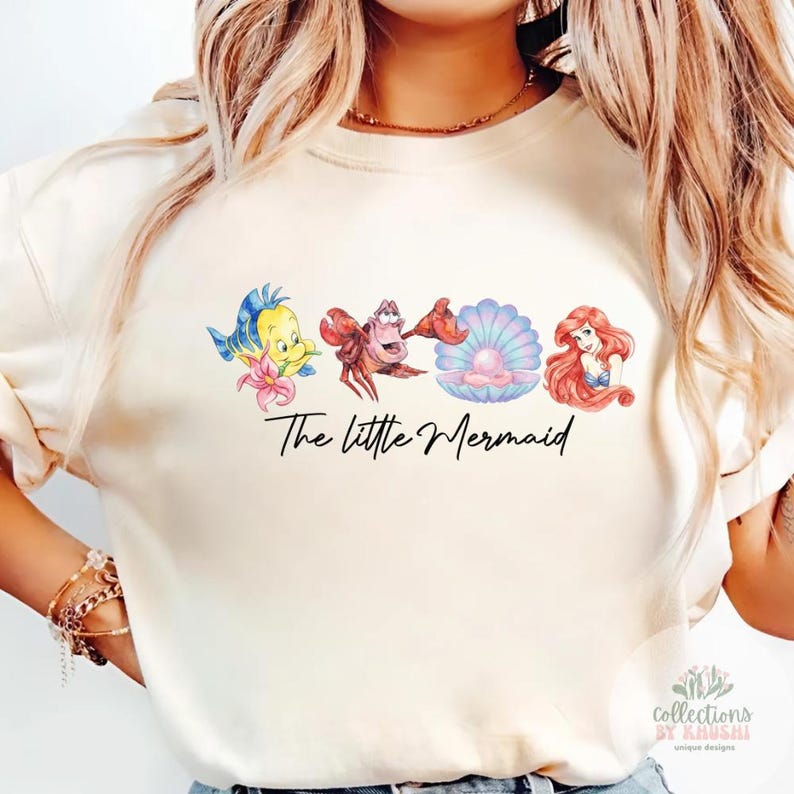 May include: A cream-colored crewneck t-shirt featuring a graphic print of Flounder, Sebastian, a pearl in a shell, and Ariel. Below the characters, the text "The little Mermaid" is printed in a black, cursive script font across the center chest.