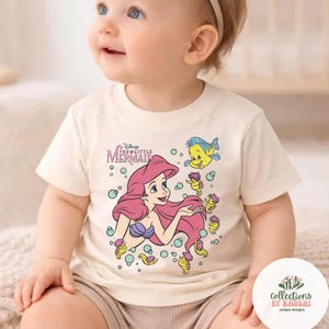 Może przedstawiać: Kremowy t-shirt z kr&oacute;tkim rękawem z nadrukiem graficznym przedstawiającym Arielkę z Małej Syrenki z długimi r&oacute;żowymi włosami, fioletowym topem z muszelek oraz kilkoma ż&oacute;łtymi konikami morskimi i niebieską rybką w jasnoniebieskich bąbelkach.
