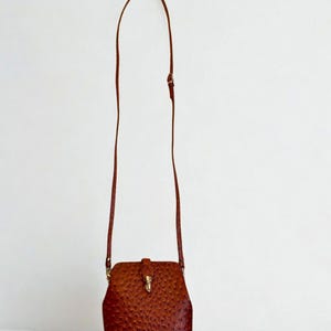 May include: A brown leather crossbody bag with a textured finish. The bag features a rectangular body with a curved top and a long, thin strap. It is secured with a flap and a gold-coloured clasp.