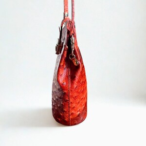 May include: A red leather handbag with a textured surface and a long strap. The bag has a rounded shape and a metal clasp closure. The bag is hanging against a white background.