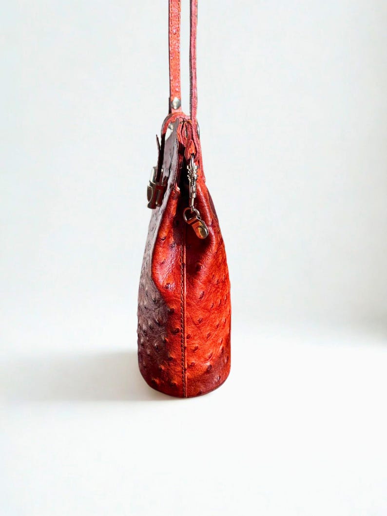 May include: A red leather handbag with a textured surface and a long strap. The bag has a rounded shape and a metal clasp closure. The bag is hanging against a white background.