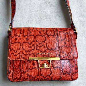 May include: A red crossbody bag featuring a snake print design. The bag has a rectangular shape with a flap closure and a gold-coloured clasp. The strap is adjustable and made of the same material as the bag. The word "SMARTBAG" is printed on the front.