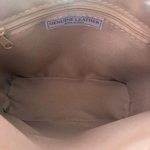 May include: Inside view of a beige leather bag with a zip closure. A rectangular label inside the bag reads "GENUINE LEATHER MADE IN ITALY". The bag's interior is a light beige colour.