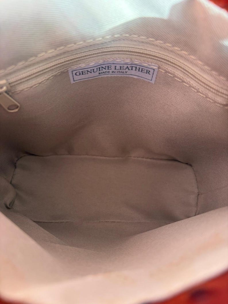 May include: Inside view of a beige leather bag with a zip closure. A rectangular label inside the bag reads "GENUINE LEATHER MADE IN ITALY". The bag's interior is a light beige colour.