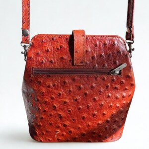 May include: A red-orange leather crossbody bag with a textured, dotted surface. The bag has a zip closure, a flap with a buckle, and a long, adjustable strap. The hardware is silver-toned.