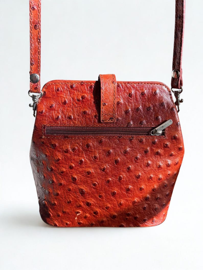 May include: A red-orange leather crossbody bag with a textured, dotted surface. The bag has a zip closure, a flap with a buckle, and a long, adjustable strap. The hardware is silver-toned.