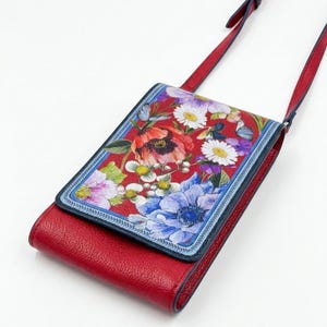 May include: Red leather crossbody bag with a floral print flap. The flap features a vibrant design of red poppies, white daisies, and blue anemones. The bag has a long red strap and a navy blue trim.