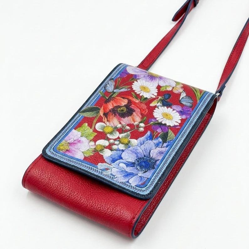 May include: Red leather crossbody bag with a floral print flap. The flap features a vibrant design of red poppies, white daisies, and blue anemones. The bag has a long red strap and a navy blue trim.