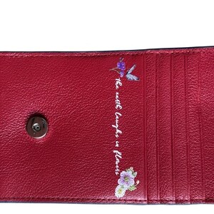 May include: A red leather wallet with a magnetic snap closure. The interior features multiple card slots. The phrase "the earth laughs in flowers" is printed in white script, accompanied by a hummingbird and floral design.
