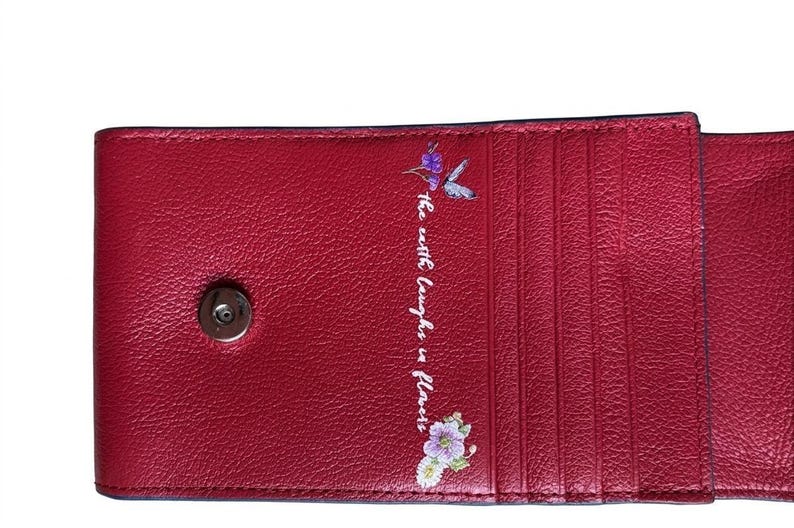 May include: A red leather wallet with a magnetic snap closure. The interior features multiple card slots. The phrase "the earth laughs in flowers" is printed in white script, accompanied by a hummingbird and floral design.