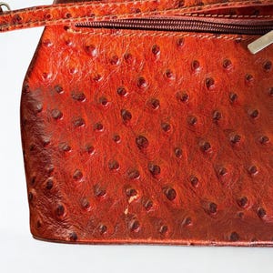 May include: A reddish-brown leather handbag with a textured surface. The bag has a zip closure and a strap. The leather has a distinctive pattern of small, raised bumps, giving it a unique appearance.