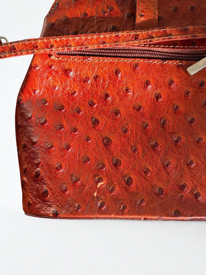 May include: A reddish-brown leather handbag with a textured surface. The bag has a zip closure and a strap. The leather has a distinctive pattern of small, raised bumps, giving it a unique appearance.