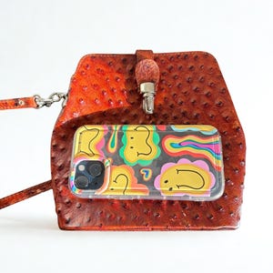 May include: A brown leather crossbody bag with a textured finish. A clear phone case with a colourful, cartoon design is visible. The bag has a strap and a metal clasp.