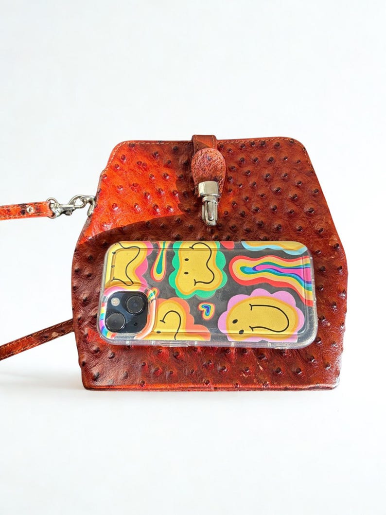 May include: A brown leather crossbody bag with a textured finish. A clear phone case with a colourful, cartoon design is visible. The bag has a strap and a metal clasp.