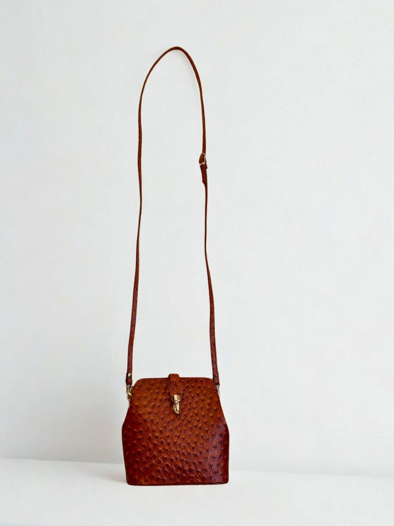 May include: A brown leather crossbody bag with a textured finish. The bag features a rectangular body with a curved top and a long, thin strap. It is secured with a flap and a gold-coloured clasp.