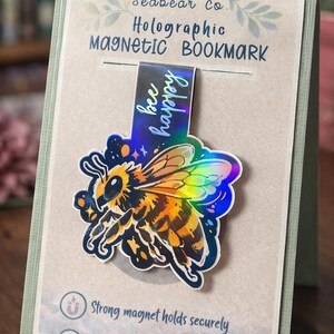 Könnte beinhalten: Holografischer Magnet-Lesezeichen mit einer bunten Bienenillustration. Das Lesezeichen trägt den Text "bee happy" und ist an einer Karte befestigt, auf der "Seabear Co. Holographic Magnetic Bookmark" steht. Auf der Karte steht auch "Strong magnet holds securely".