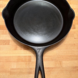 May include: A black cast iron skillet with a long handle and a rounded cooking surface. The number 7 is embossed on the handle. The skillet is sitting on a wooden surface.