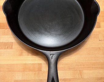 May include: A black cast iron skillet with a long handle and a rounded cooking surface. The number 7 is embossed on the handle. The skillet is sitting on a wooden surface.