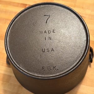 Könnte beinhalten: Ein schwarzer gusseiserner Topf mit der Zahl 7 auf dem Deckel. Die Worte "MADE IN USA" und die Initialen "F.B.K." sind ebenfalls sichtbar. Der Topf hat zwei Griffe und eine abgerundete Form.