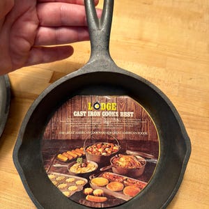 Puede incluir: Una pequeña sartén de hierro fundido negro con un asa. La sartén tiene una superficie de cocción circular y un asa con un agujero para colgar. El texto en el inserto dice "Lodge Cast Iron Cooks Best".