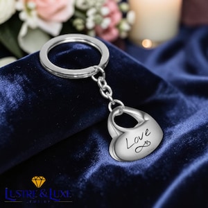 Puede incluir: Llavero plateado en forma de corazón con la palabra "Love" en escritura cursiva. El llavero tiene un llavero y una cadena. El fondo es de tela de terciopelo azul oscuro.