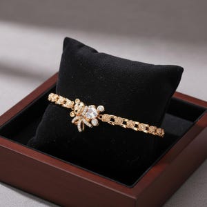 May include: Gold-toned bracelet featuring a teddy bear charm with a bow and sparkling accents. The bracelet has a delicate chain design with rose-shaped links. The bracelet is displayed on a black velvet cushion inside a wooden box.