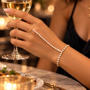 May include: A gold-toned hand chain bracelet featuring a delicate chain connecting a ring to a wrist bracelet, both adorned with a continuous row of small, clear, round-cut sparkling stones that catch the light, creating a refined accessory.